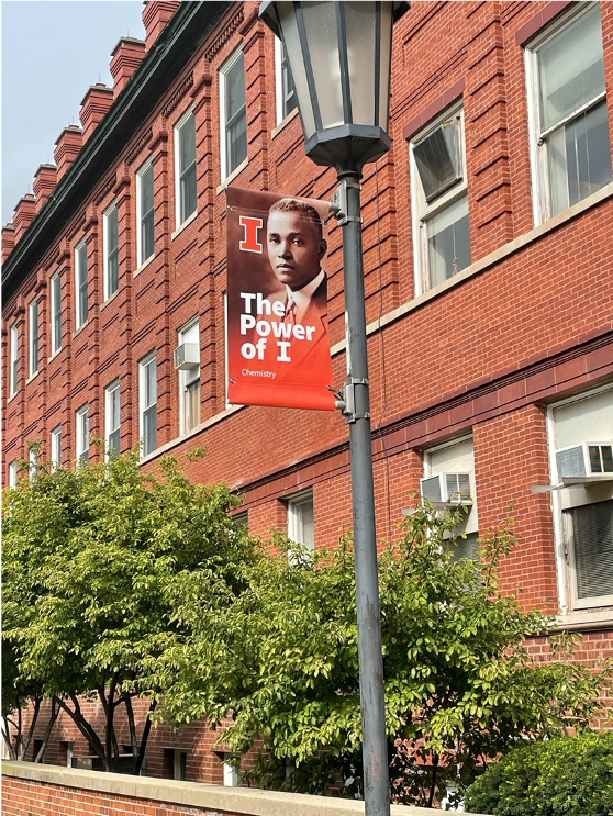 Orange banner on a lamp post in front of a brick building. The banner shows a portrait of young Black man with text "The Power of I"