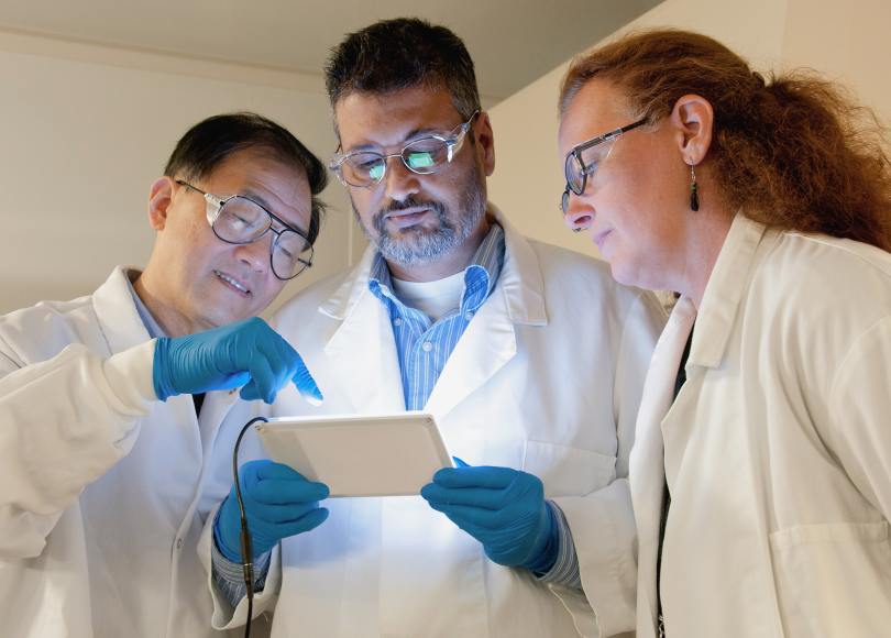 three people in lab coats and safety goggles look down on a glowing tablet